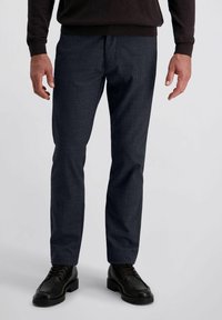 Man in een donker geruit broek, zwarte veterschoenen en een donker langemouwen shirt, staande tegen een effen lichte achtergrond.