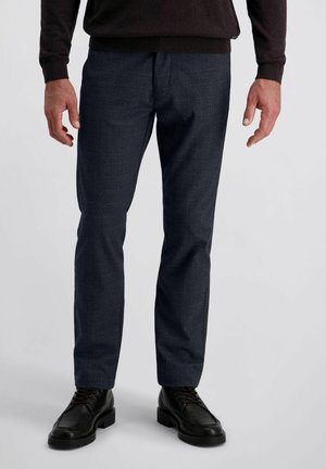 Man in een donker geruit broek, zwarte veterschoenen en een donker langemouwen shirt, staande tegen een effen lichte achtergrond.