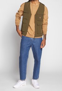 Chaleco sin mangas verde oliva con múltiples bolsillos, llevado sobre un sudadera color beige y jeans azules. Zapatillas blancas completan el conjunto.
