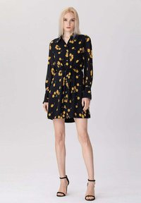 Femme aux longs cheveux blonds portant une robe noire à boutons avec des motifs de fruits jaunes et des sandales à talons noirs, se tenant sur un fond blanc.