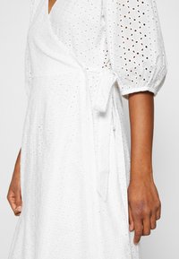 Bílý zavinovací šaty s nabíranými rukávy, zdobené výšivkou s dírkami, s vázáním v pase a splývavou siluetou. Hladká bavlněná textura.