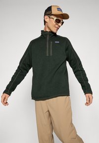 Mørkegrønn fleece-pullover med halv-zip krage, kontrasterende sømmer, og Patagonia-logo. Brukt med khaki-bukser og en caps.