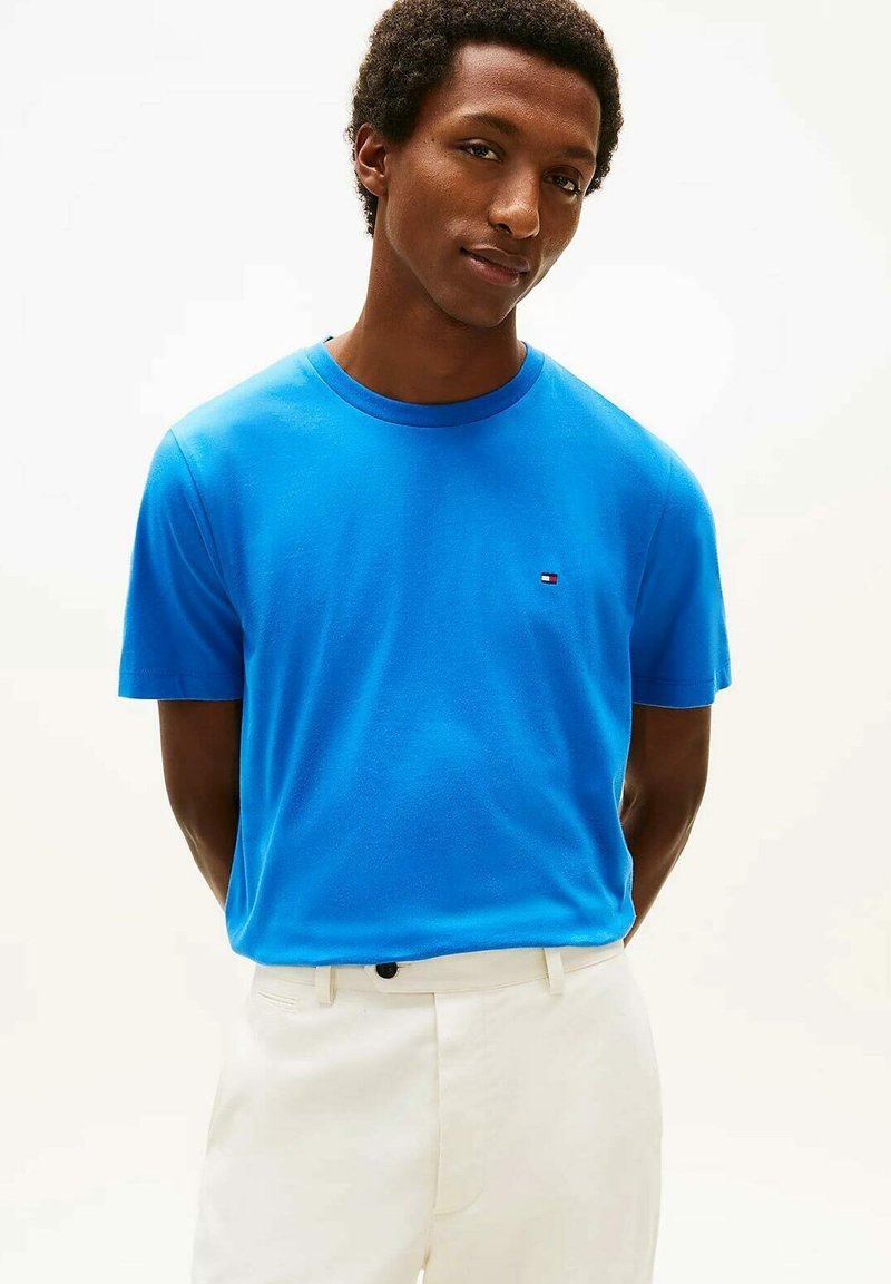 Homme portant un t-shirt à manches courtes bleu vif avec un petit logo sur la poitrine et un pantalon de couleur claire, debout devant un fond uni.