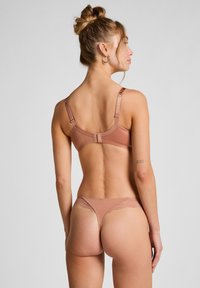 Vrouw met een knot in haar haar, gekleed in een taupe bh en bijpassende thong met kant, die van zich af kijkt tegen een effen lichtgrijze achtergrond.