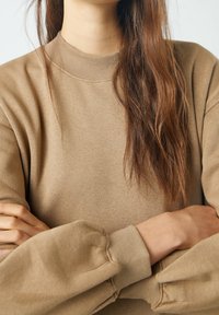 Osoba z długimi brązowymi włosami, ubrana w beżowy sweter z bufiastego rękawami, z założonymi na siebie rękami, na jednolitym jasnym tle.