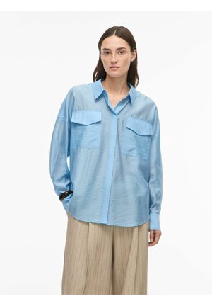 Kvinne iført en oversized lyseblå skjorte med brystlommer og beige bukser med vid ben, stående med én hånd i lommen.