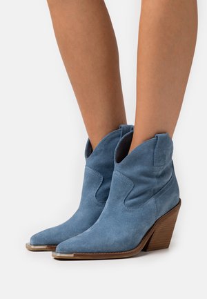 Botas de tobillo de ante azul con punta afilada y parte superior curva. Cuentan con un tacón de bloque de madera y costuras decorativas en los lados.