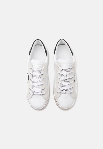Paire de baskets basses blanches avec des touches noires et des lacets blancs imprimés d'un texte en écriture cursive noire sur un fond clair uni.