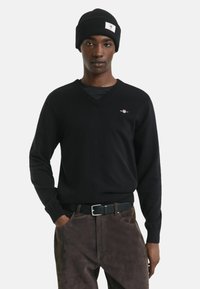 Maglione di lana nera con scollo a V e logo piccolo. Abbinato a un berretto nero a costine e pantaloni marrone scuro, dotati di cintura.