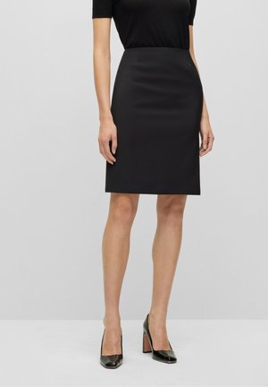 Kvinde iført en tætsiddende sort knælængde blyantskørt med en sort top og sorte høje hæle, stående på en ensfarvet baggrund.