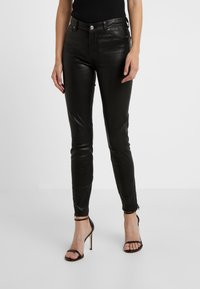 Kobieta w dopasowanych czarnych skórzanych spodniach z suwakami na kostkach i czarnych sandałach na wysokim obcasie, stojąca na tle jednolitego białego tła.