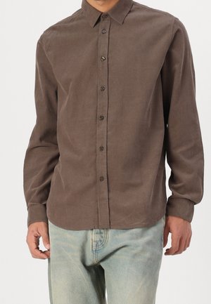 Homme portant une chemise marron à manches longues boutonnée et un jean bleu clair, debout, bras détendus le long du corps, devant un fond uni.