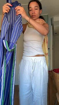 Woman holding a blue, white, and green striped garment while wearing a white tank top, white pants, and a patterned scarf indoors.