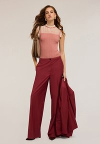 Top rosa a coste senza maniche con scollatura trasparente, abbinato a pantaloni larghi bordeaux. Il modello tiene una borsa marrone e una giacca rossa.