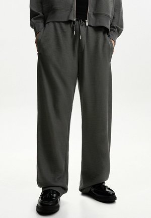 Pantalons à texture gaufrée grise avec une coupe ample, dotés de poches latérales et d'une taille élastique avec cordon de serrage. Accompagnés de chaussures noires.