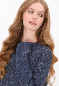 Gebreide trui in donkergroen met lichtgroene spikkels, geribbelde manchetten en een ronde halslijn. Zachte textuur voegt warmte en diepte toe aan het ontwerp.