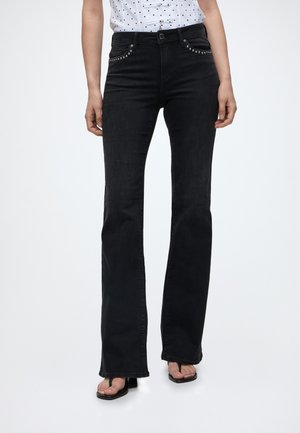 Kvinde iført sorte højttaljede flare jeans med nittede lommer, kombineret med sorte åbne hælede sandaler og en hvid skjorte med prikker.