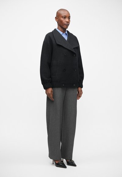 Abrigo de lana negro tipo peacoat con cierre de doble botonadura y bolsillos laterales, combinado con pantalones de pierna ancha en cuadros grises y tacones negros de punta.
