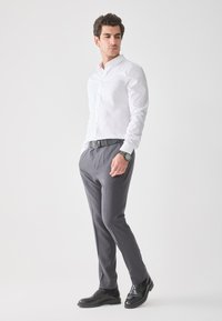 White button-up shirt, gray trousers, black belt, and black shoes. Model stands with a neutral expression against a light background.
