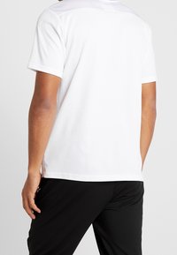 Valkoinen urheilupaita lyhyillä hihoilla, teksturoitu kangas ja hieman kaareva helma. Mustat housut rentoa istuvuutta.