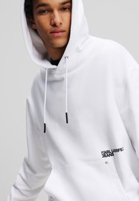 Weißer Kapuzenpullover mit Kordelzügen, ausgestattet mit einer großen Fronttasche und schwarzem "KARL LAGERFELD JEANS" Logo auf der unteren linken Seite.