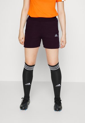 Persoon draagt zwarte Adidas sportshorts, zwarte Adidas kniekousen met witte strepen, zwarte voetbalschoenen en een oranje shirt, staand op een grijze vloer.
