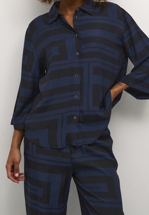 Femme portant une chemise boutonnée bleu marine et noire à motif géométrique et un pantalon assorti, une main dans la poche et l'autre détendue sur le côté.