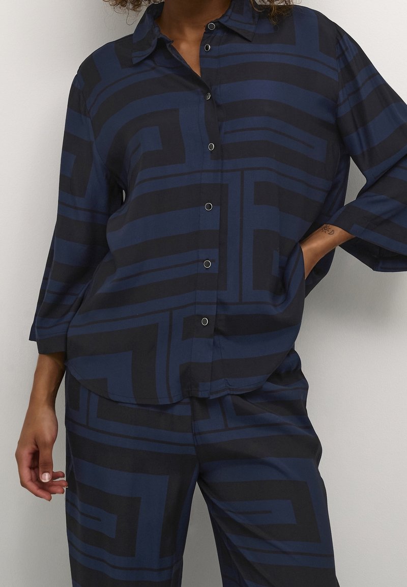 Femme portant une chemise boutonnée bleu marine et noire à motif géométrique et un pantalon assorti, une main dans la poche et l'autre détendue sur le côté.