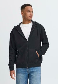 Svart zip-up hettegenser med snor, store frontlommer og ribbede mansjetter, båret over en hvit t-skjorte og blå jeans.