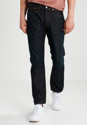 Mies, joka pukeutunut tummansinisiin suora lahkeisiin farkkuihin ja valkoisiin tennareihin, seisoo yksinkertaisen vaalean taustan edessä.