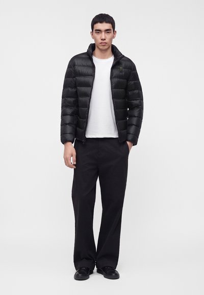 Jeune homme debout, faisant face à l'objectif, portant une doudoune noire sur un t-shirt blanc, un pantalon large noir et des chaussures noires sur un fond uni.