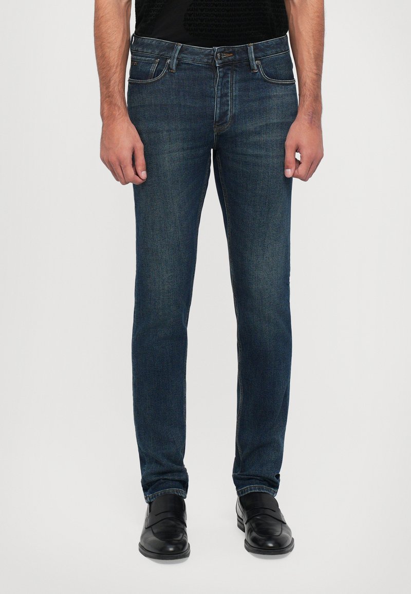 Jeans de ganga azul-escuro com um corte ajustado, cinco bolsos, leve desgaste e detalhes de costura contrastantes. Usados com sapatos pretos.