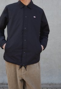 Person wearing a black button-up jacket with a logo on the chest and beige corduroy pants, standing against a plain gray wall.