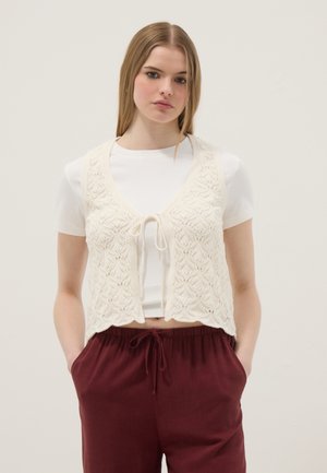 Mujer joven que lleva un chaleco de punto texturizado blanco atado al frente sobre una camiseta blanca y pantalones burdeos con cordón, manos en los bolsillos.