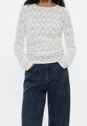 Kobieta ubrana w biały, długi koronkowy top z rękawami i ciemnoniebieskie jeansy z wysokim stanem, z rękami w kieszeniach na jednolitym tle.