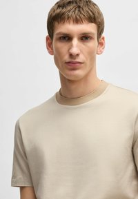 T-shirt beige à manches courtes fabriqué dans un tissu texturé. Il présente un col rond avec une bordure contrastante de couleur marron clair. Design simple.