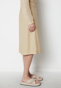 Beige Baumwollrock mit ausgestelltem Design, knielang, mit zwei seitlichen Taschen und sichtbarem Knopfdetail; kombiniert mit hellbraunen Slipper-Sandalen.