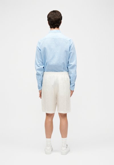 Άνδρας που στέκεται με την πλάτη προς τα εμπρός, φορώντας ανοιχτό μπλε μακρύ μανίκι πουκάμισο, λευκά σορτς, λευκές κάλτσες και λευκά αθλητικά παπούτσια σε απλό φόντο.