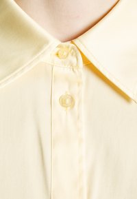 Gros plan d'une chemise jaune pâle à col montrant deux boutons et une couture soignée autour du col et de la patte de boutonnage.
