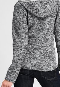 Persona che indossa un hoodie grigio e nero mescolato e jeans blu scuro, mostrata di spalle con un braccio leggermente piegato.