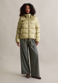 Puffige, hellgrüne Jacke mit Reißverschluss und Kordelzugkapuze, kombiniert mit karierten, weit geschnittenen Hosen und braunen Stiefeletten.