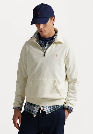 Crème pullover sweatshirt met een kwart-rits, voorzak en klein logo. Gedragen over een blauw geruit overhemd, gecombineerd met marineblauwe broek en een marineblauwe pet.