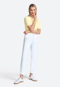 Haut tricot jaune à manches courtes, jeans blancs taille haute et baskets claires. La tenue présente une coupe décontractée et une texture subtile dans les matériaux.