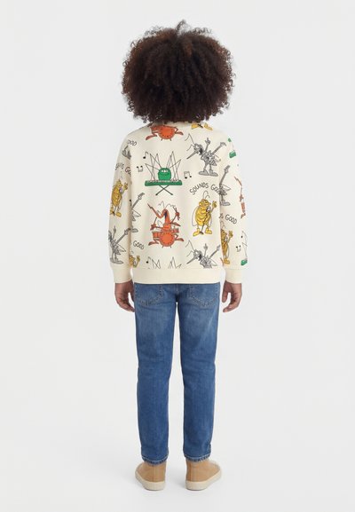 Vaikas su banguotais plaukais dėvi kreminės spalvos megztinį su spalvotais animaciniais muzikos instrumentų ir personažų raštais, mėlynas džinsas ir rudos spalvos batus.