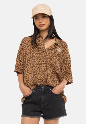 Femme portant une casquette beige Von Dutch, une chemise ample à imprimé léopard avec boutons, et un short en jean noir, les mains dans les poches.