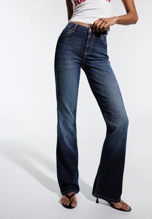 Femme portant un jean flare taille haute bleu foncé et des talons noirs à brides, tenant la ceinture du jean d'une main.