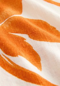 Gros plan sur un tissu blanc avec des formes courbes abstraites orange et un tissage texturé visible.