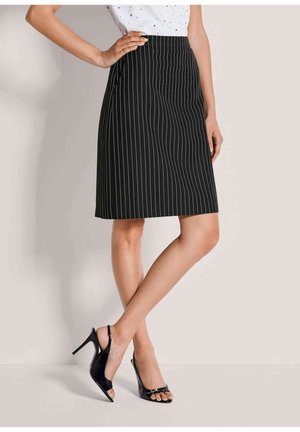 Frau trägt kniehohen schwarzen Nadelstreifenrock und schwarze offene High Heels, posiert mit einer Hand in der Hüfte vor schlichtem Hintergrund.