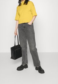 Osoba v žlutém tričku s krátkým rukávem, šedých džínách, černých silných mokasínách, držící černou tašku na rameno, s jednou rukou v kapse.