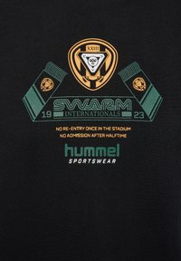 Czarna bawełniana koszulka z nadrukowanymi grafikami w kolorze zielonym i pomarańczowym. Design zawiera sportowe motywy oraz tekst: "SWARM INTERNATIONALS 2023", "hummel SPORTSWEAR."